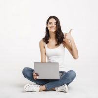 Portrait of a happy girl dressed in tank-top holding laptop while sitting on the floor and showing thumbs up gesture isolated over white background